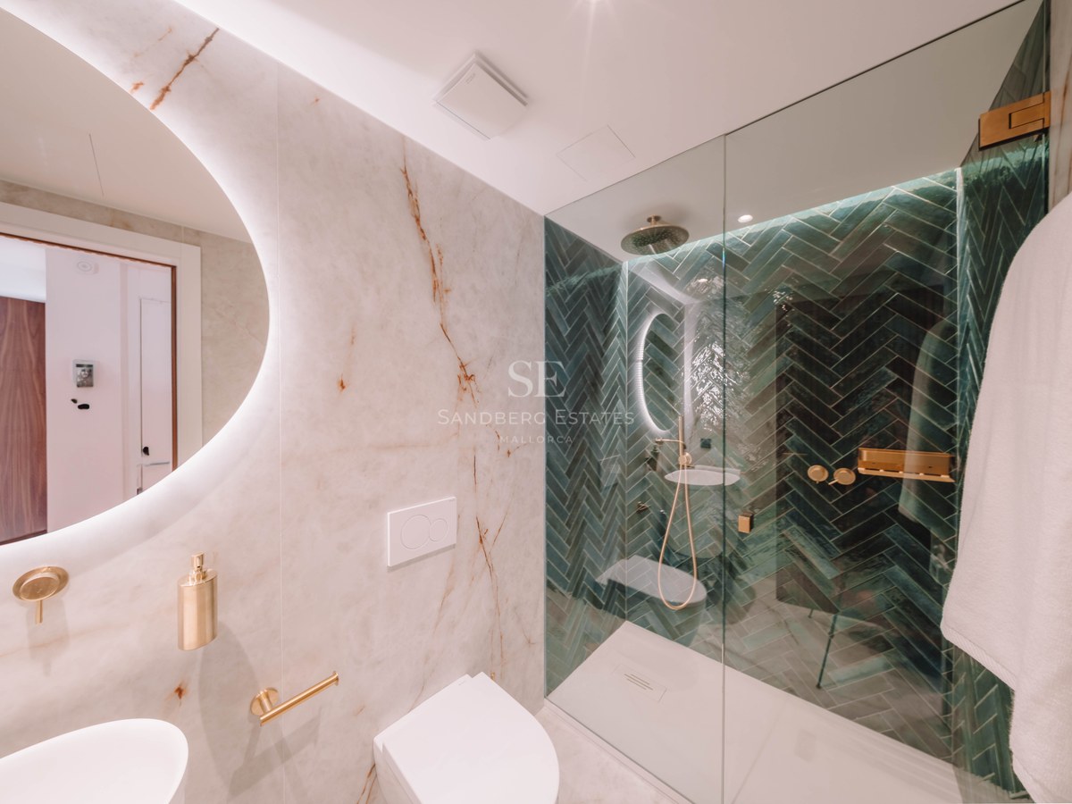 Modern bathroom with white marble walls, a round backlit mirror, and a walk-in shower with green herringbone tiles.