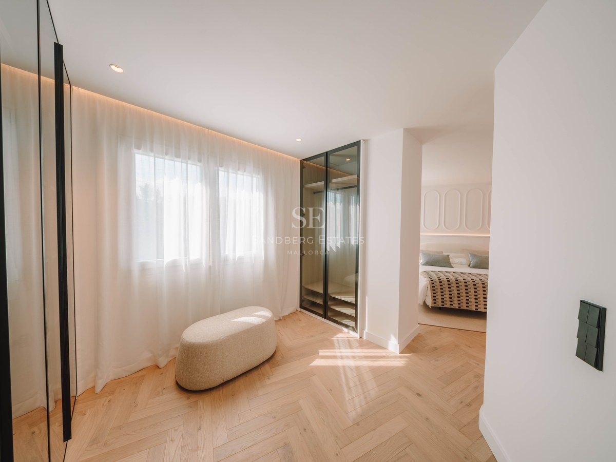 Bright dressing area with herringbone wood flooring, a beige ottoman, and glass wardrobe leading to a bedroom.