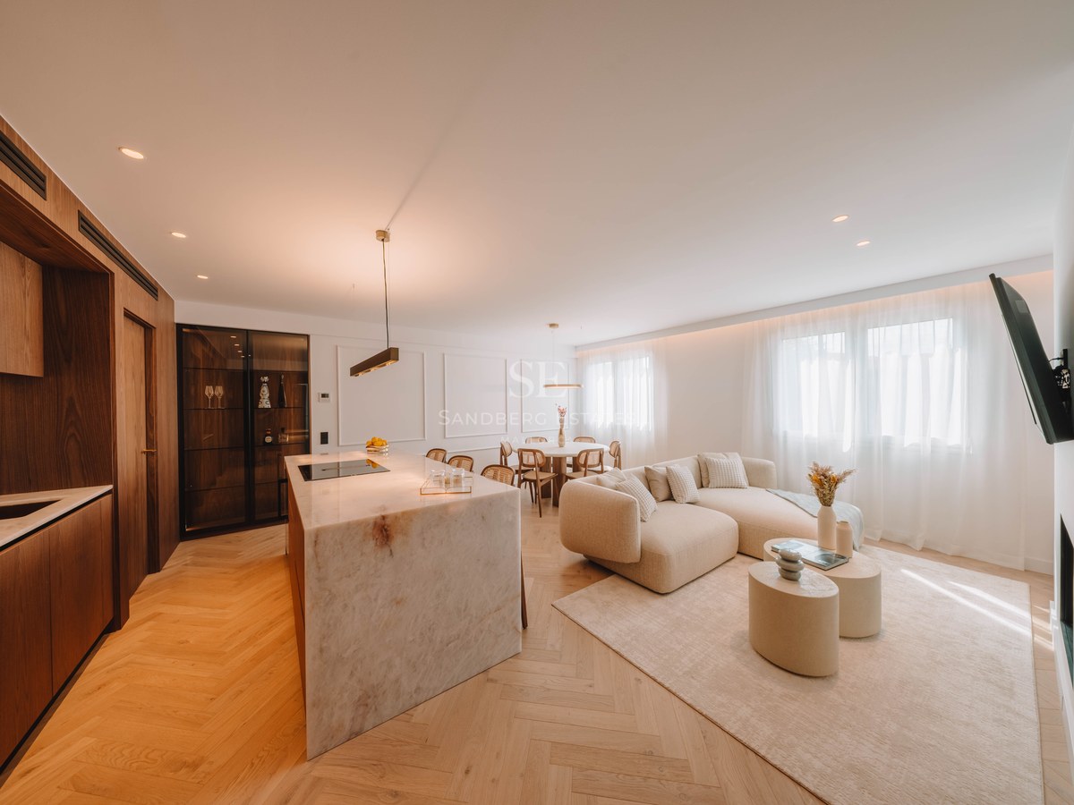 Modern open-plan living room with marble kitchen island, beige sectional sofa, and light wood herringbone floors.