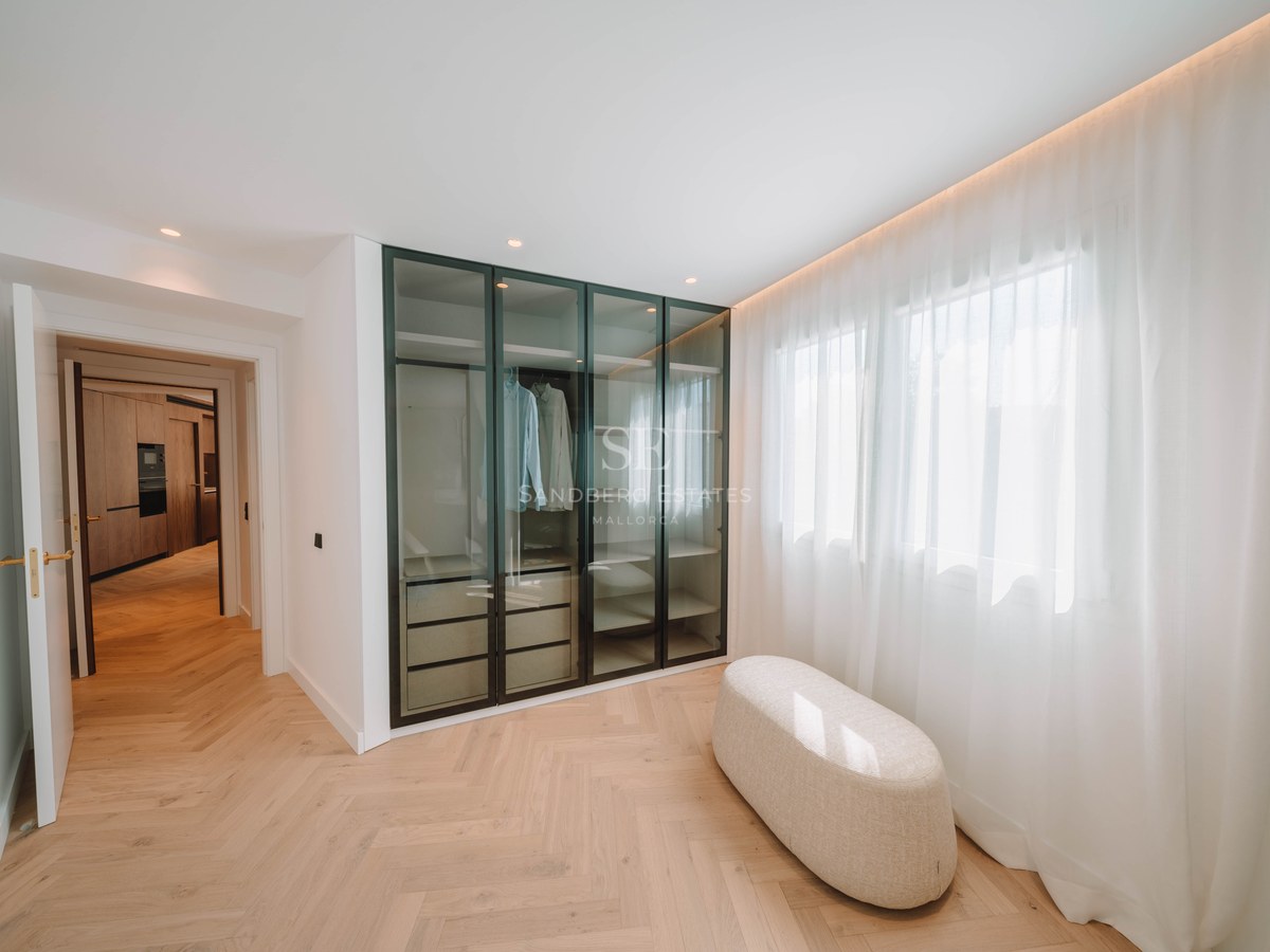Modern walk-in closet with herringbone wood floors, glass wardrobe, and a central ottoman next to a window.