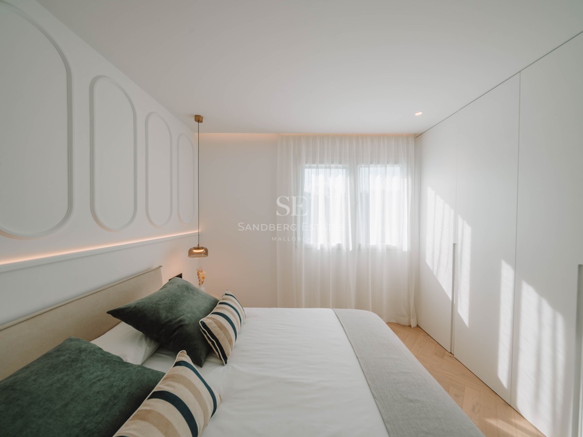 Bright bedroom with white paneled walls, a double bed with green pillows, herringbone wood floors, and built-in closets.