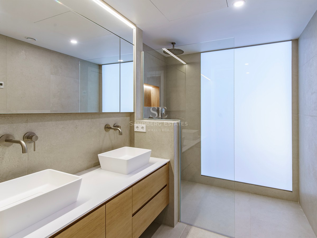 Minimalist bathroom with double white sinks on a wooden vanity, walk-in shower, and large backlit mirror.