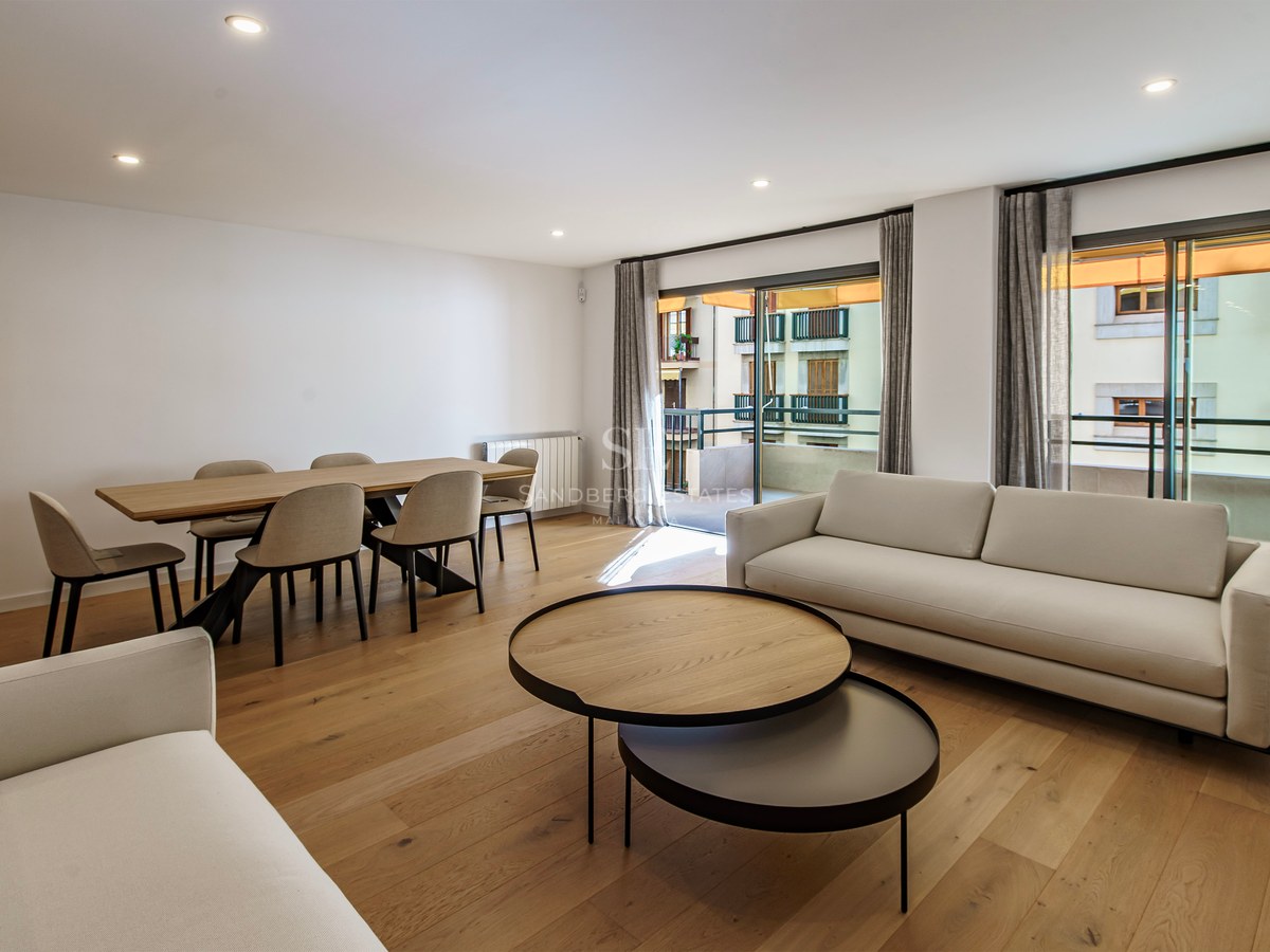 Modern living room featuring white sofas, wooden coffee tables, a dining set, and large windows leading to a balcony.