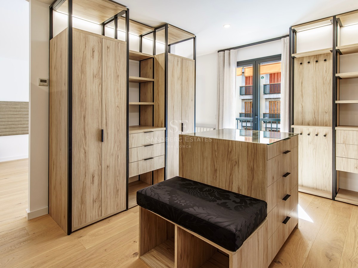 Spacious walk-in closet featuring light wood cabinetry, a central island with a glass top, and a black cushioned bench.