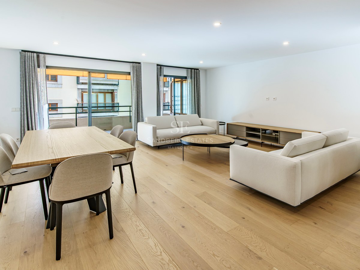 Open-plan living room with white sofas, wooden dining table, and large windows leading to a balcony.