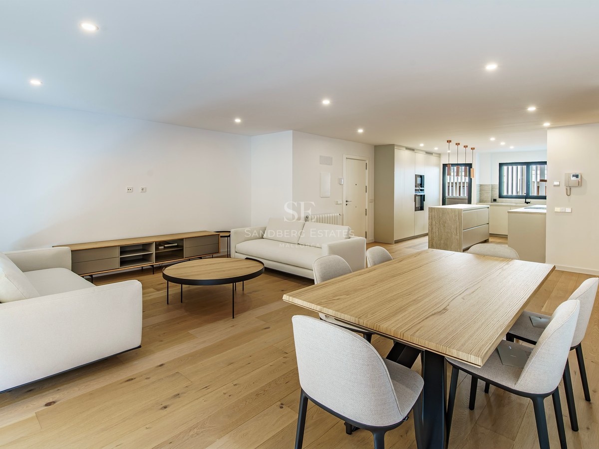 Spacious modern room featuring white sofas, a large wooden dining table, and a contemporary kitchen in the background.