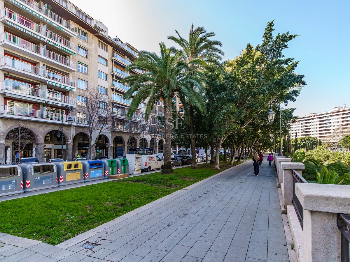 Paved city promenade with palm trees next to a multi-story stone building with balconies under a blue sky.