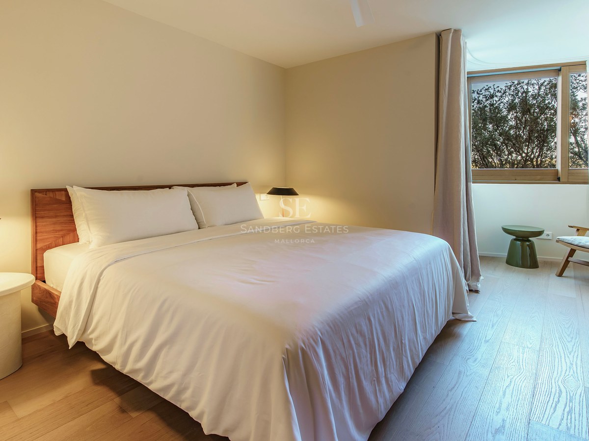 Modernes Schlafzimmer mit weißer Bettwäsche, Holzkopfteil, Designerkleuchten und einem Sessel am Fenster.
