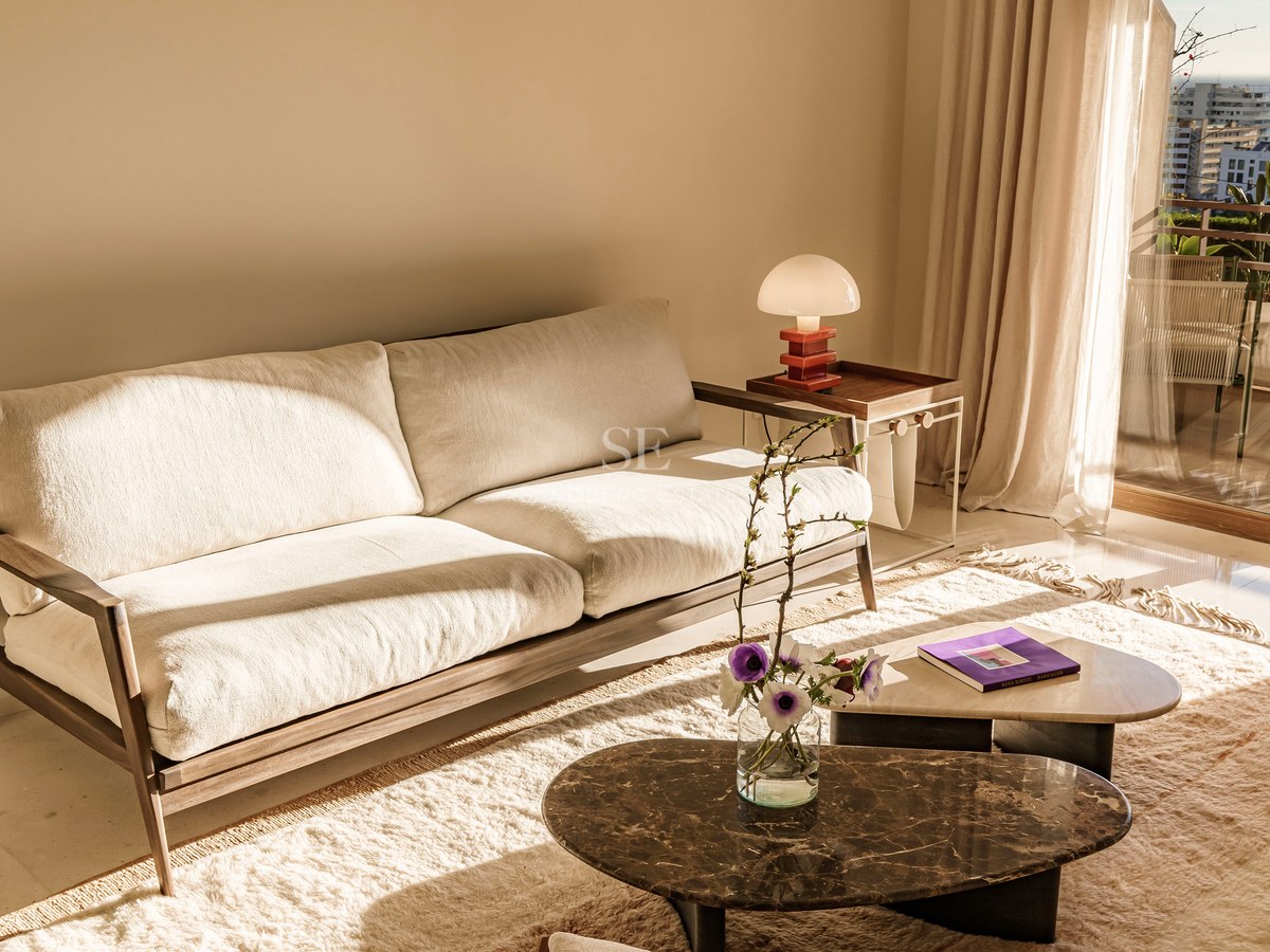 Cremefarbenes Sofa und Marmortische in einem sonnigen Wohnzimmer mit Balkon und Meerblick.