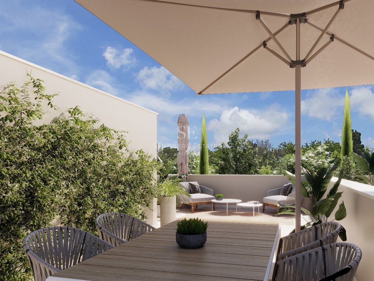 Bright outdoor terrace with a wooden dining table, large sun umbrella, lounge chairs, and lush green plants.