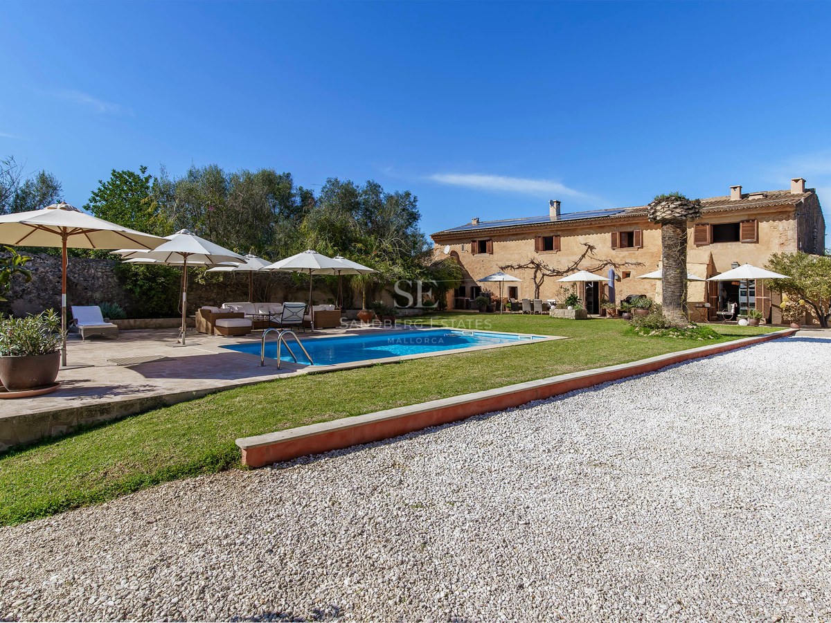Große Steinvilla mit blauem Pool, weißen Sonnenschirmen und Garten unter klarem Himmel.