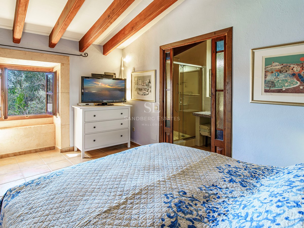Schlafzimmer mit Holzbalken, blau gemustertem Bett, sichtbarem Bad und Steinfenster mit Ausblick.