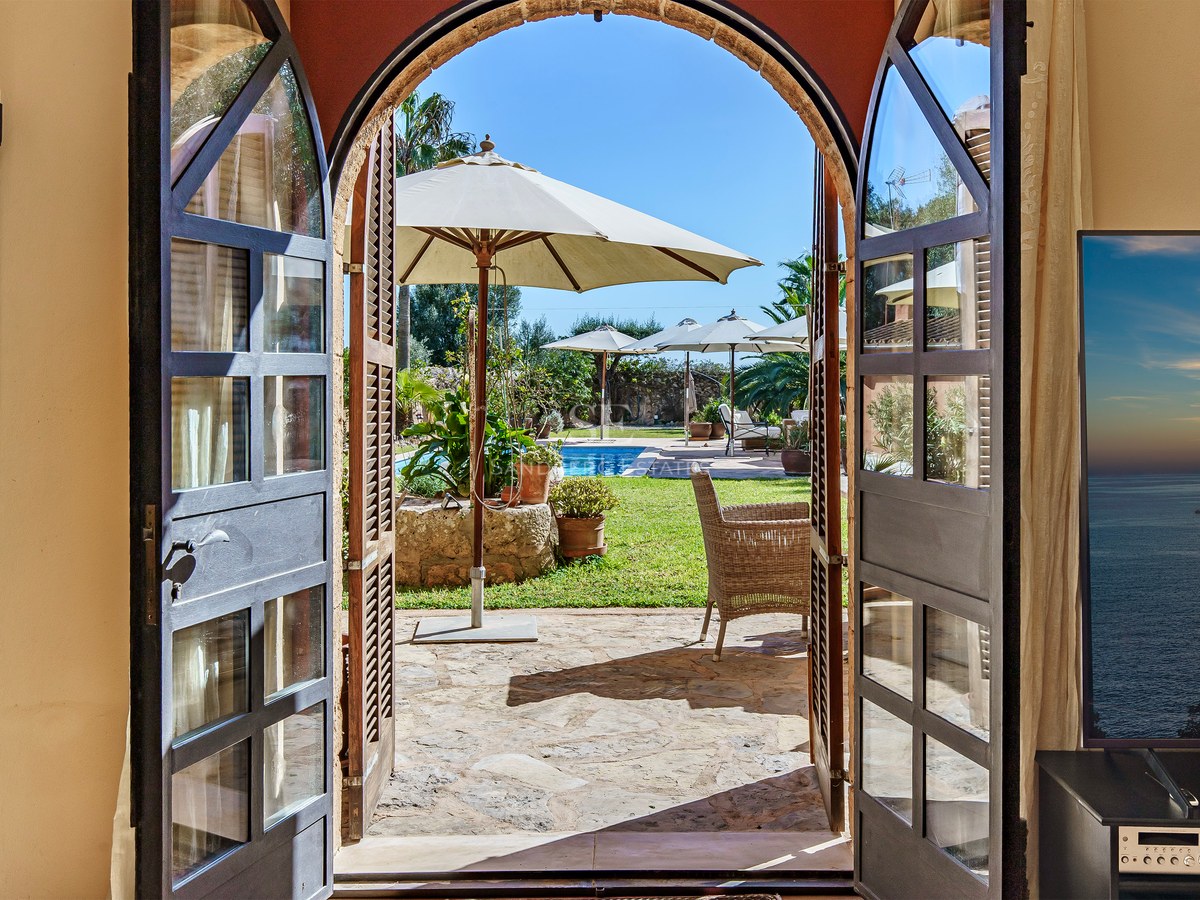 Blick durch offene Rundbogentüren auf einen privaten Pool, Garten und weiße Sonnenschirme unter blauem Himmel.