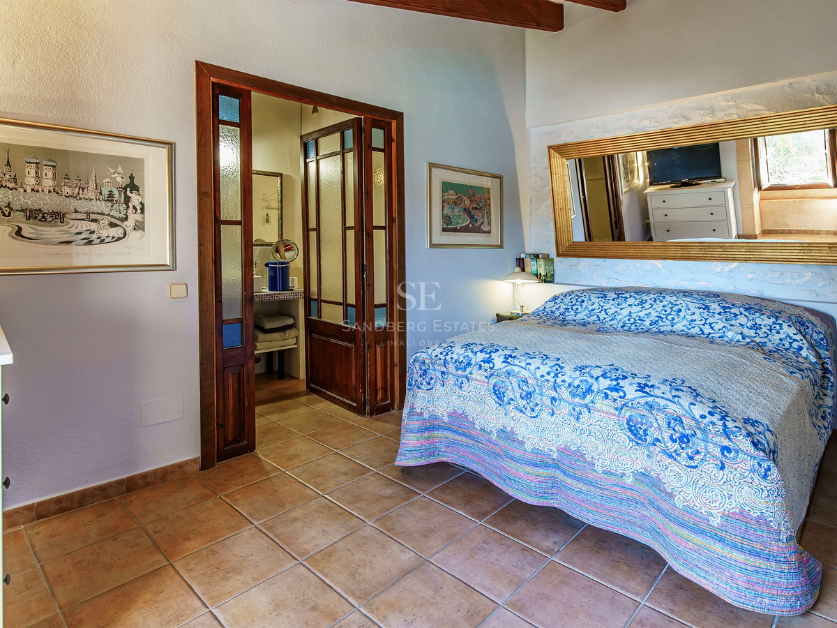Schlafzimmer mit blau gemusterter Decke, Terrakottaboden, großem Goldspiegel und Holztüren zum Bad.