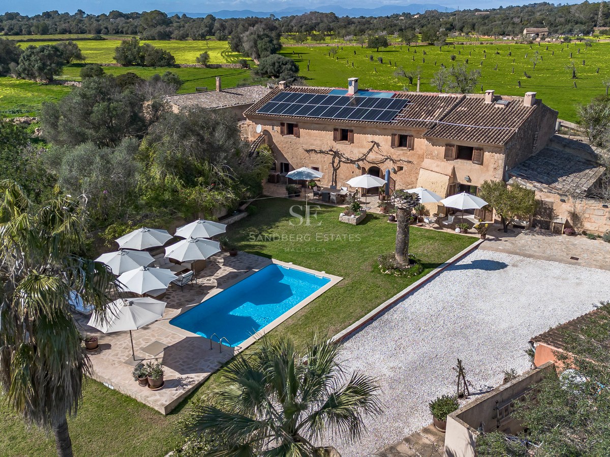 Luftaufnahme einer Steinvilla mit blauem Pool, Sonnenkollektoren und weißen Sonnenschirmen, umgeben von Feldern.