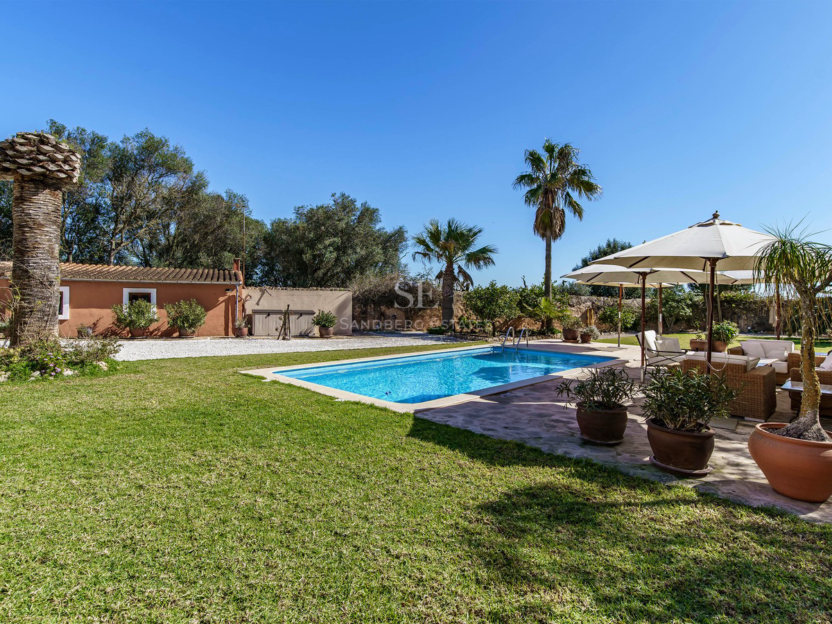Sonniger Pool umgeben von grünem Rasen, Palmen und einem Loungebereich mit Sonnenschirmen unter blauem Himmel.