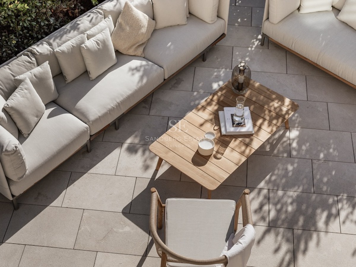 High-angle view of a luxury terrace with beige sofas, a wooden coffee table, and stone paving in dappled sunlight.