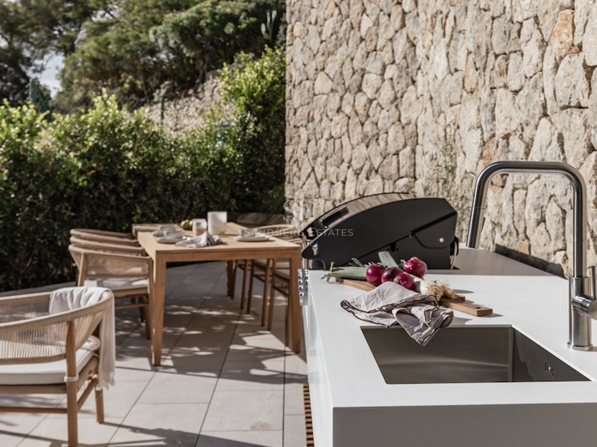 Modern outdoor kitchen with white countertop, sink, and grill next to a stone wall and wooden dining set.