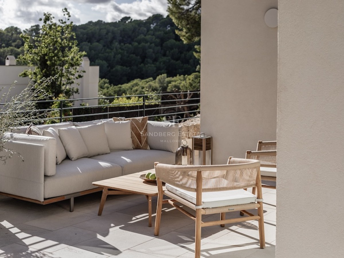 Modern outdoor seating area on a terrace with a white sofa, wooden chairs, and views of a lush green forest.