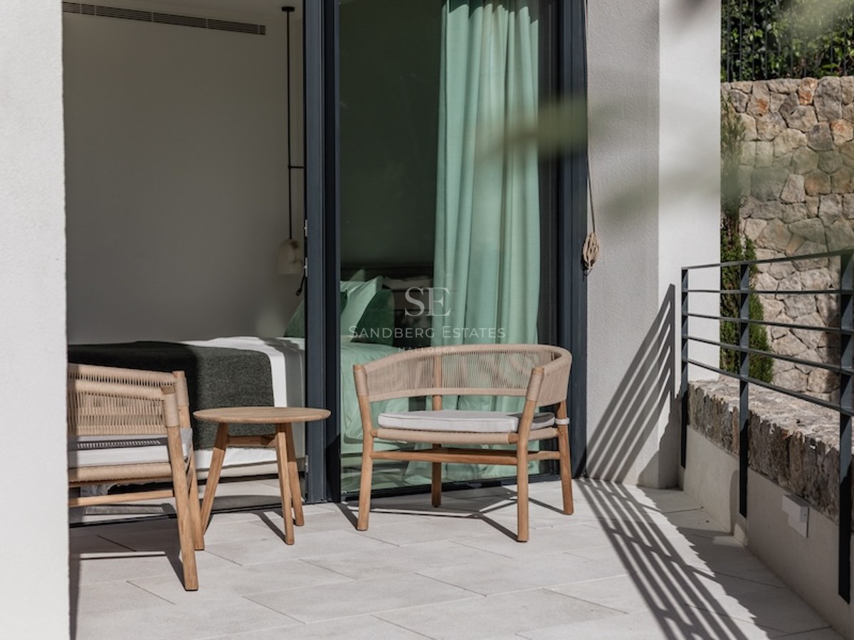 Modern terrace with woven armchairs and a small wooden table, adjacent to a bedroom with large glass sliding doors.
