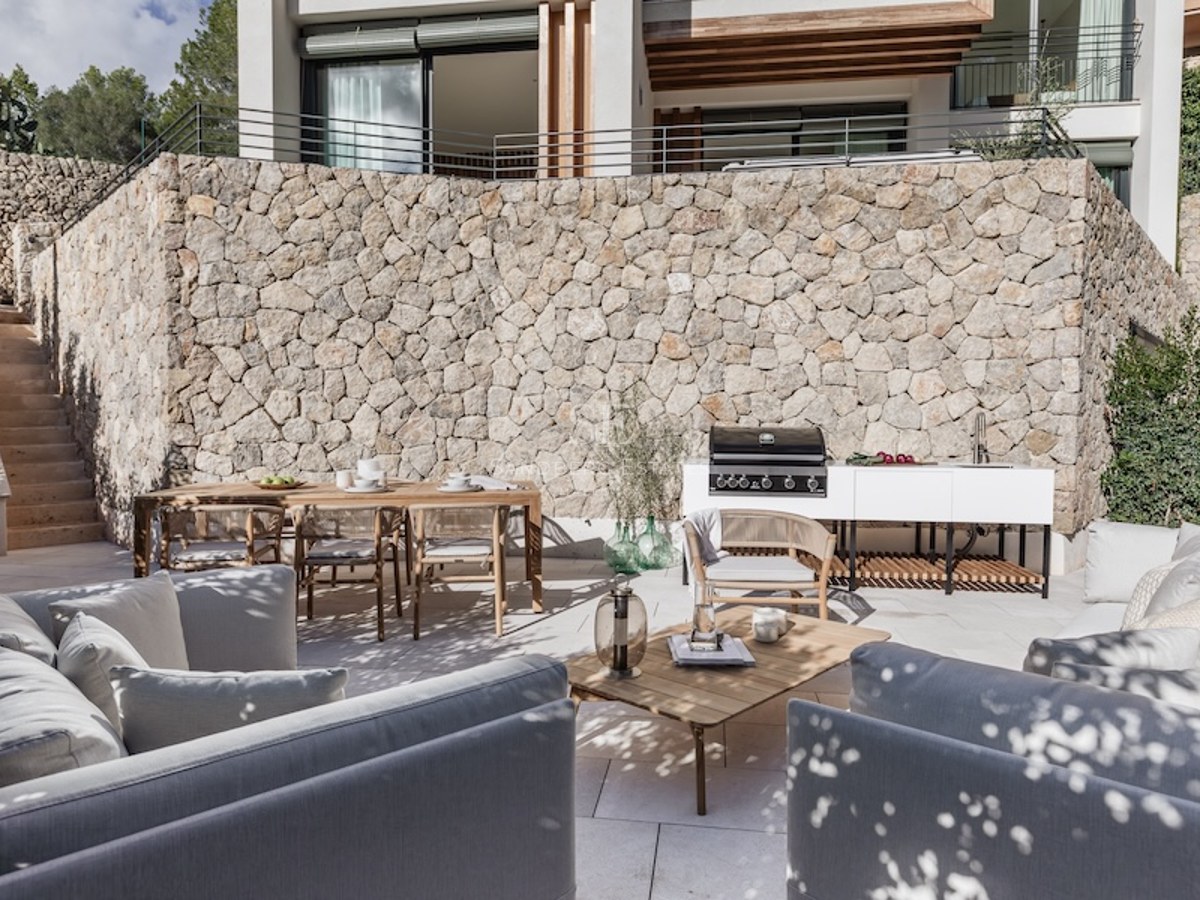 Modern terrace with a stone wall, outdoor kitchen, wooden dining table, and grey sofas under dappled natural light.