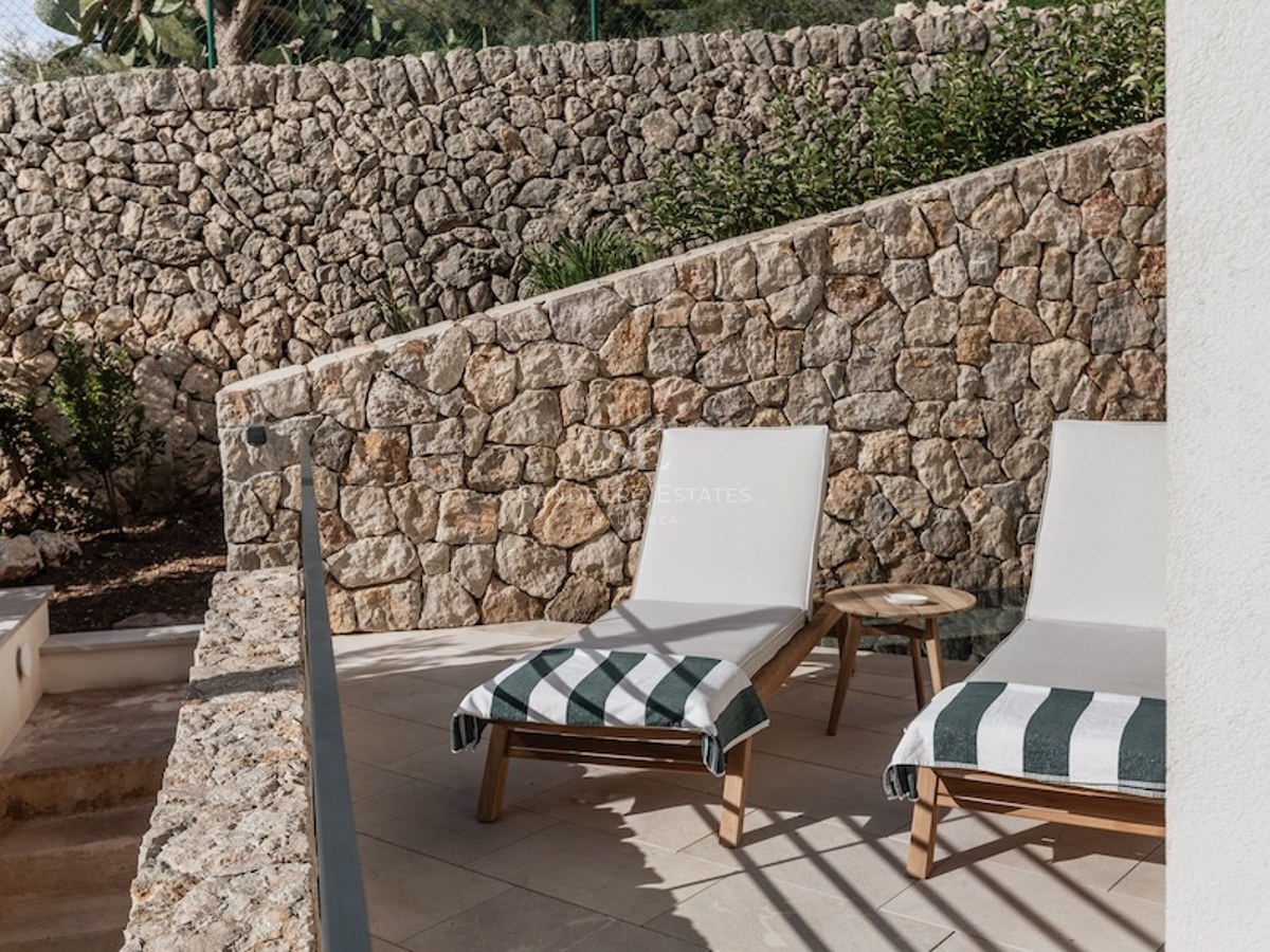 Two wooden sun loungers with white cushions in front of a rustic stone wall on a sunny terrace.
