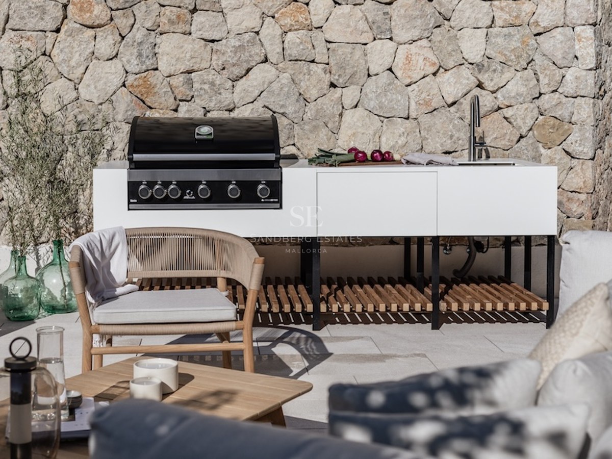 Modern white outdoor kitchen with gas grill and sink against a stone wall with designer patio furniture.