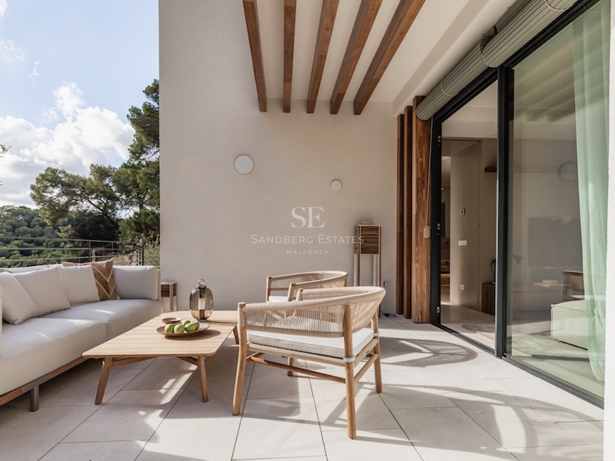 A sunlit modern terrace with a white sofa, wooden furniture, and views of lush green trees through glass doors.