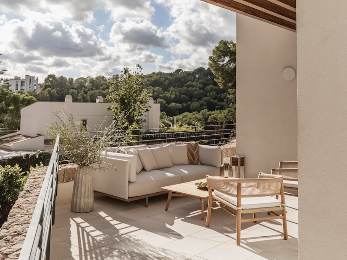 Outdoor terrace with modern sofa, wooden table, and woven chairs overlooking a lush green valley.