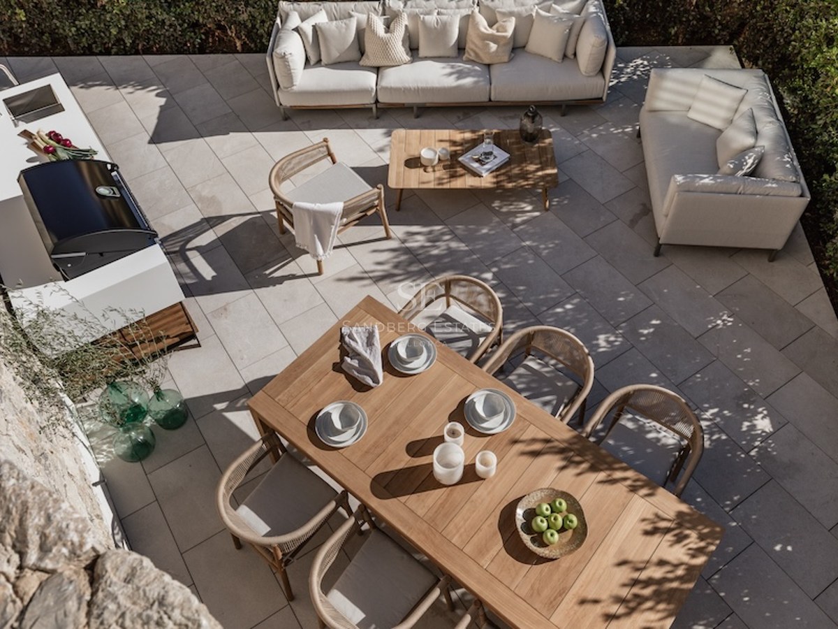Overhead view of a modern stone patio featuring a teak dining set, white sofas, and a sleek outdoor kitchen with BBQ.