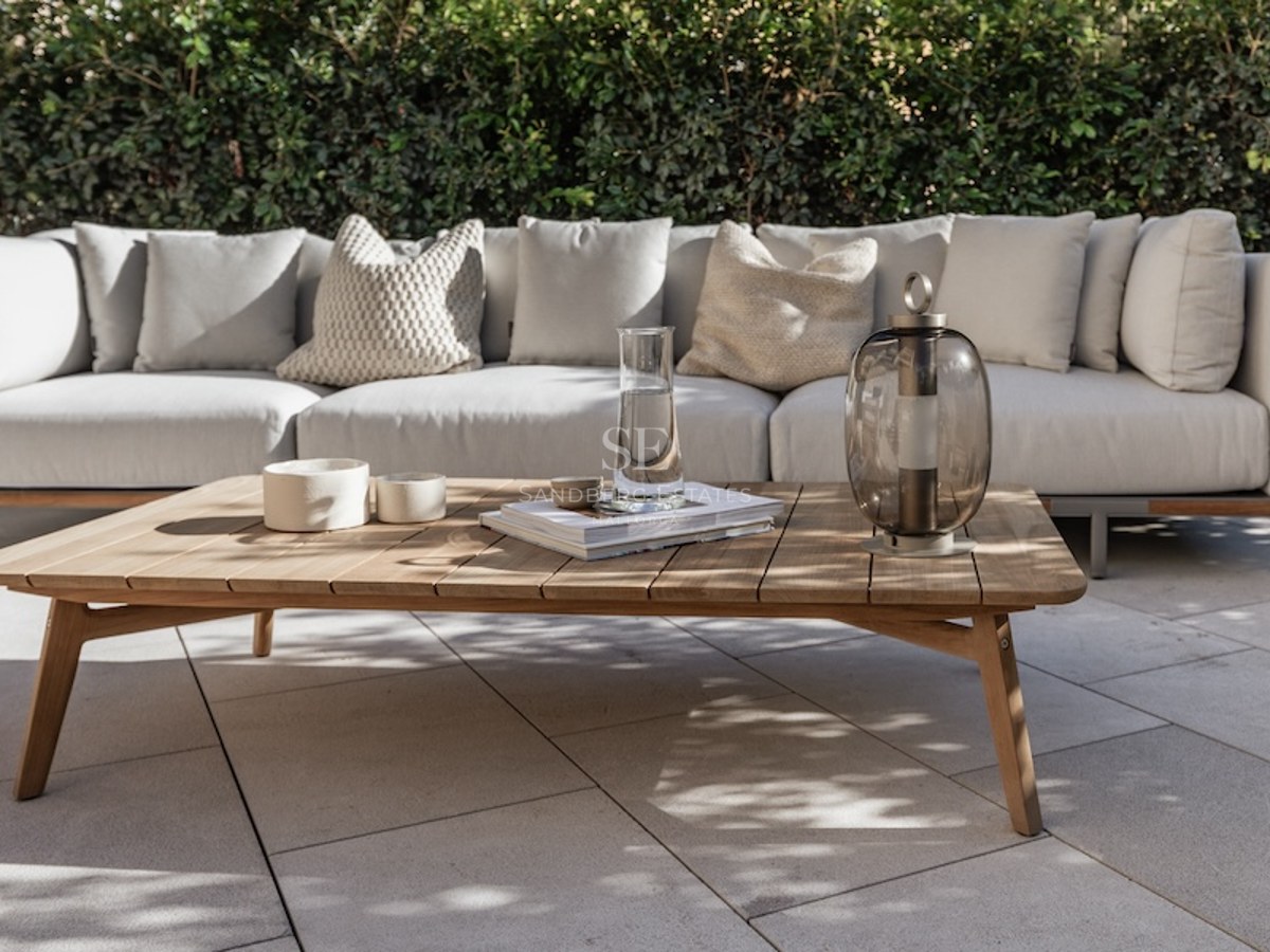 Outdoor sofa with beige cushions and a wooden coffee table on a stone patio against a green hedge.