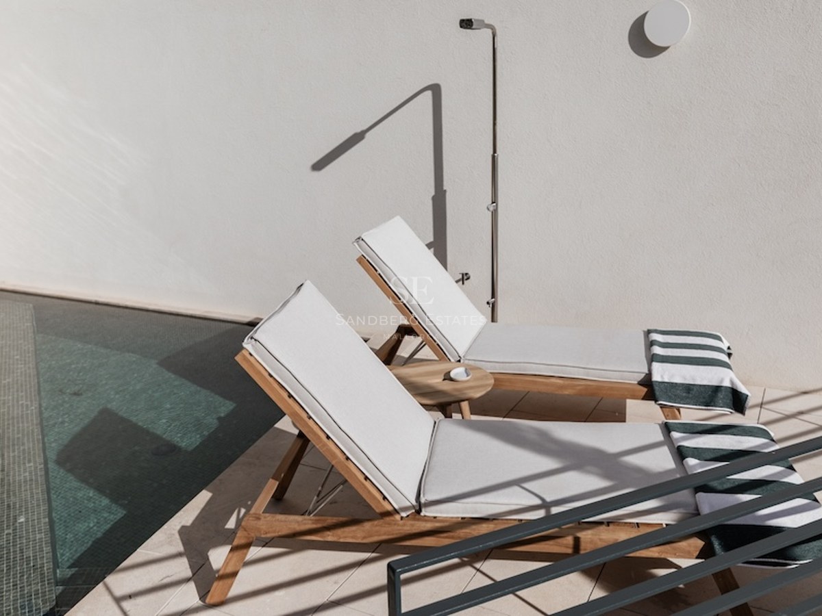 Two wooden sun loungers with white cushions next to a pool and outdoor shower on a sunny terrace.