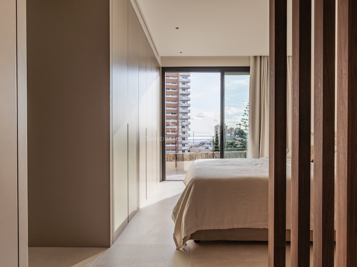 Modern bedroom with large built-in wardrobes, a bed with light linens, and a balcony window showing city buildings.