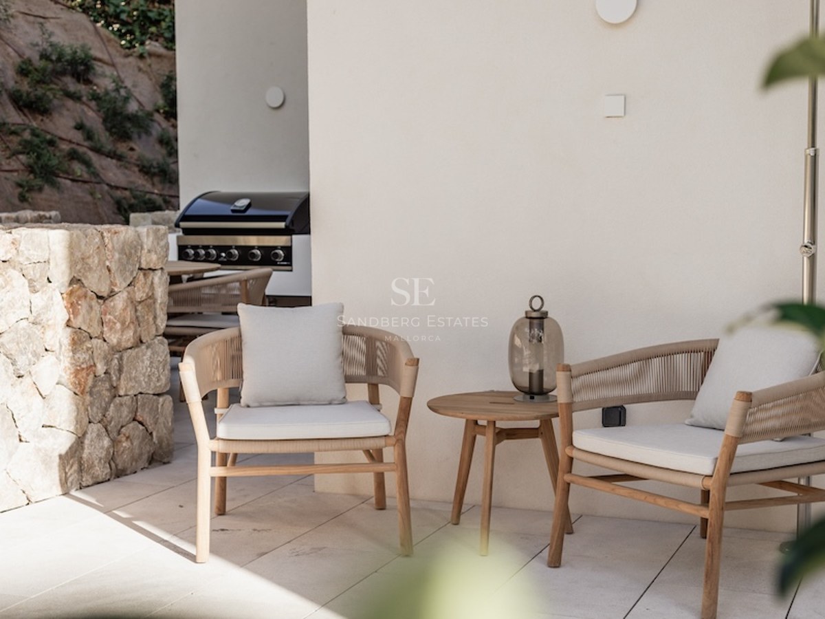 Two elegant modern armchairs with cushions next to a wooden side table on a stone terrace with a grill in the background.