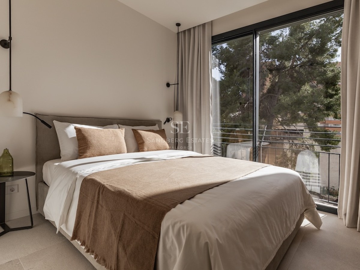 Bright bedroom with a large bed, neutral linens, designer pendant lights, and a floor-to-ceiling window viewing trees.