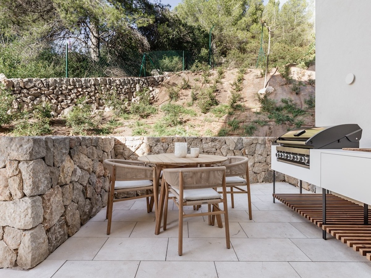 Outdoor terrace featuring a wooden dining set, stone walls, and a modern built-in grill area against a backdrop of trees.