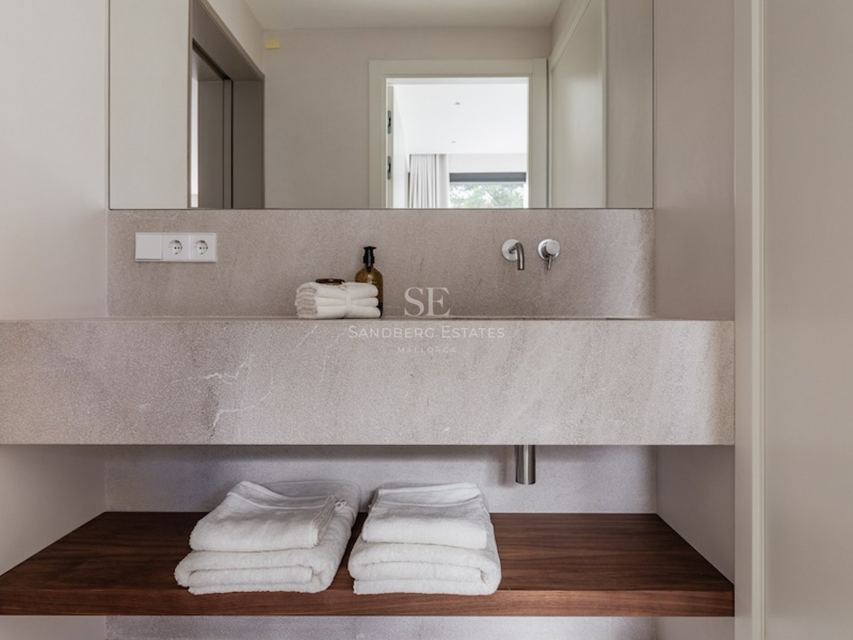 Minimalistisches Steinwaschbecken mit Wandarmatur, großem Spiegel und Holzregal mit weißen Handtüchern.