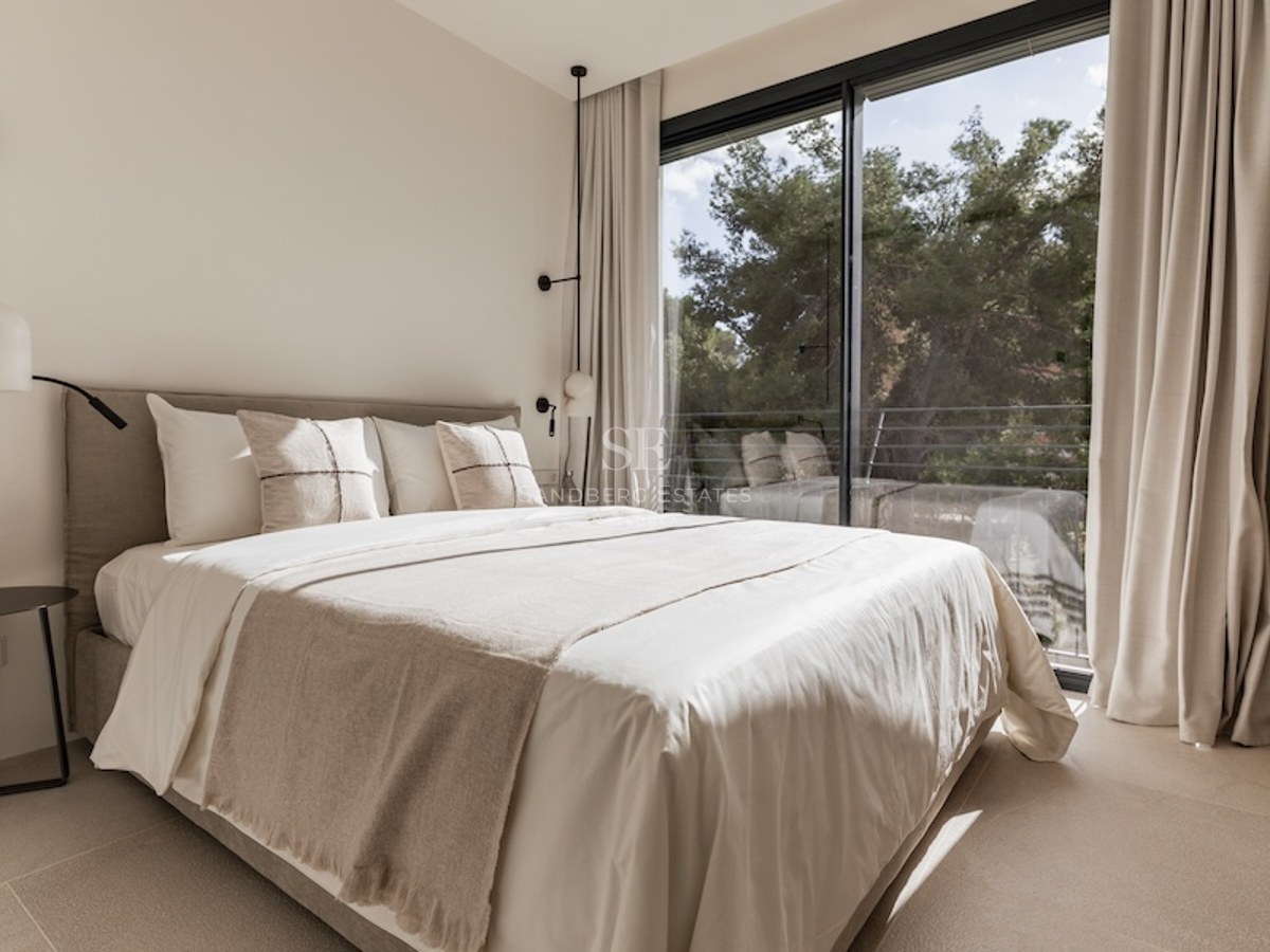 Helles Schlafzimmer mit Bett in neutralen Tönen vor einem großen Fenster mit Blick auf Bäume.