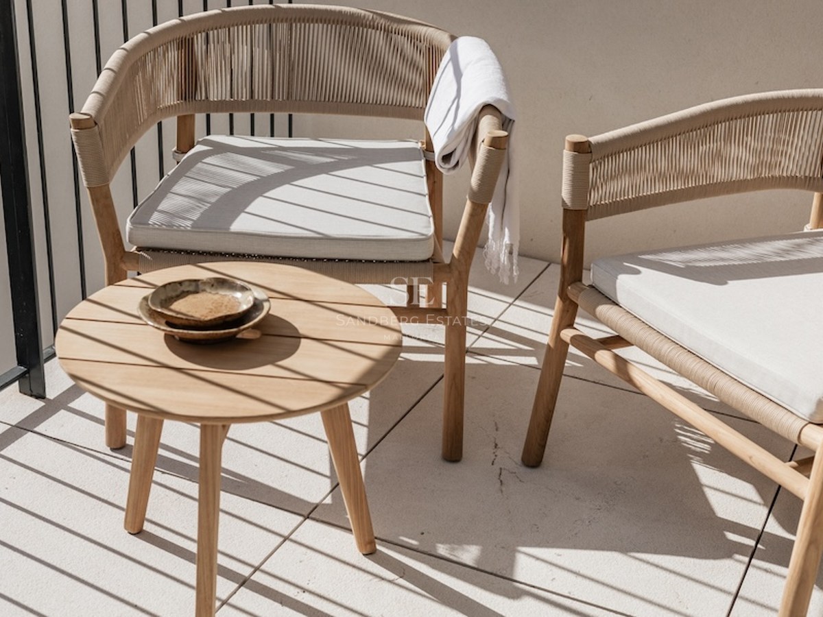 Zwei elegante geflochtene Stühle und ein runder Holzbeistelltisch auf einem sonnigen Balkon mit Schatten.