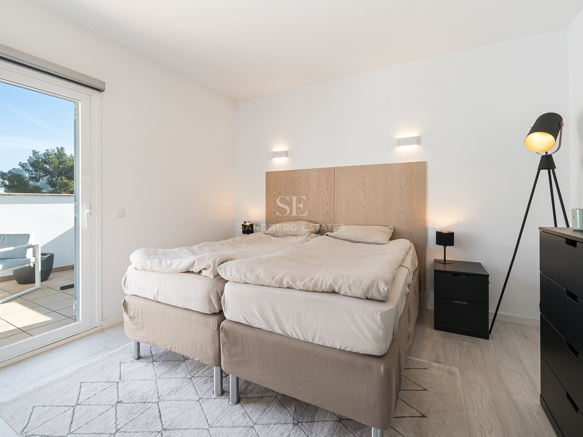 Bright bedroom featuring a double bed, light wood headboard, and a sliding glass door opening to a sunny balcony.