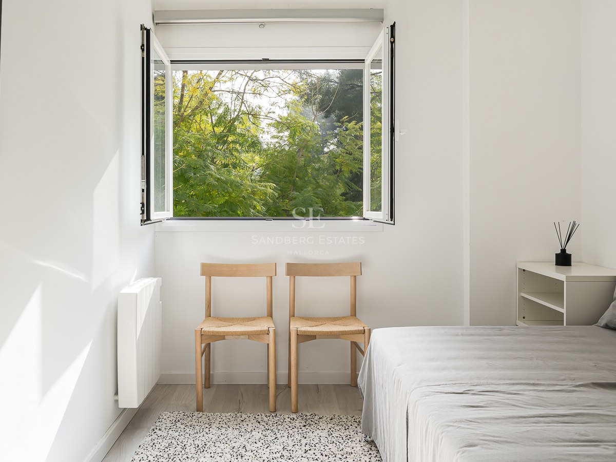 Bright minimalist bedroom with white walls, wooden chairs, grey bed, and an open window viewing lush green trees.
