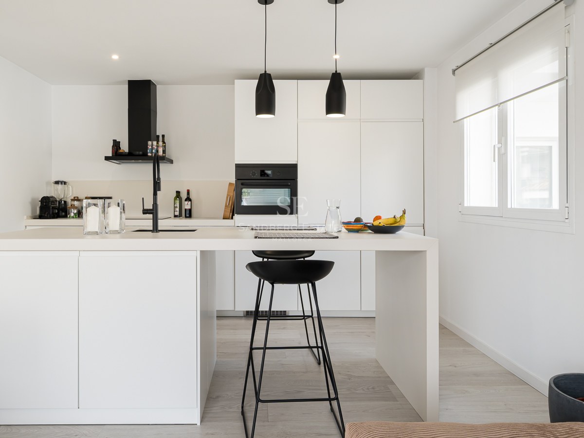 Bright white kitchen featuring a large central island with black bar stools and black hanging pendant lights.