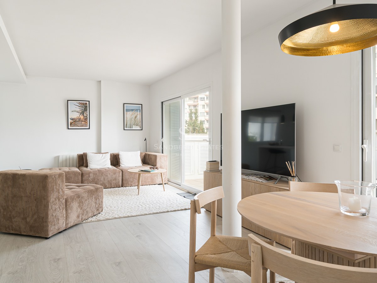 A sunlit living room featuring a brown modular sofa, wooden dining table, and large glass sliding doors leading to a balcony.