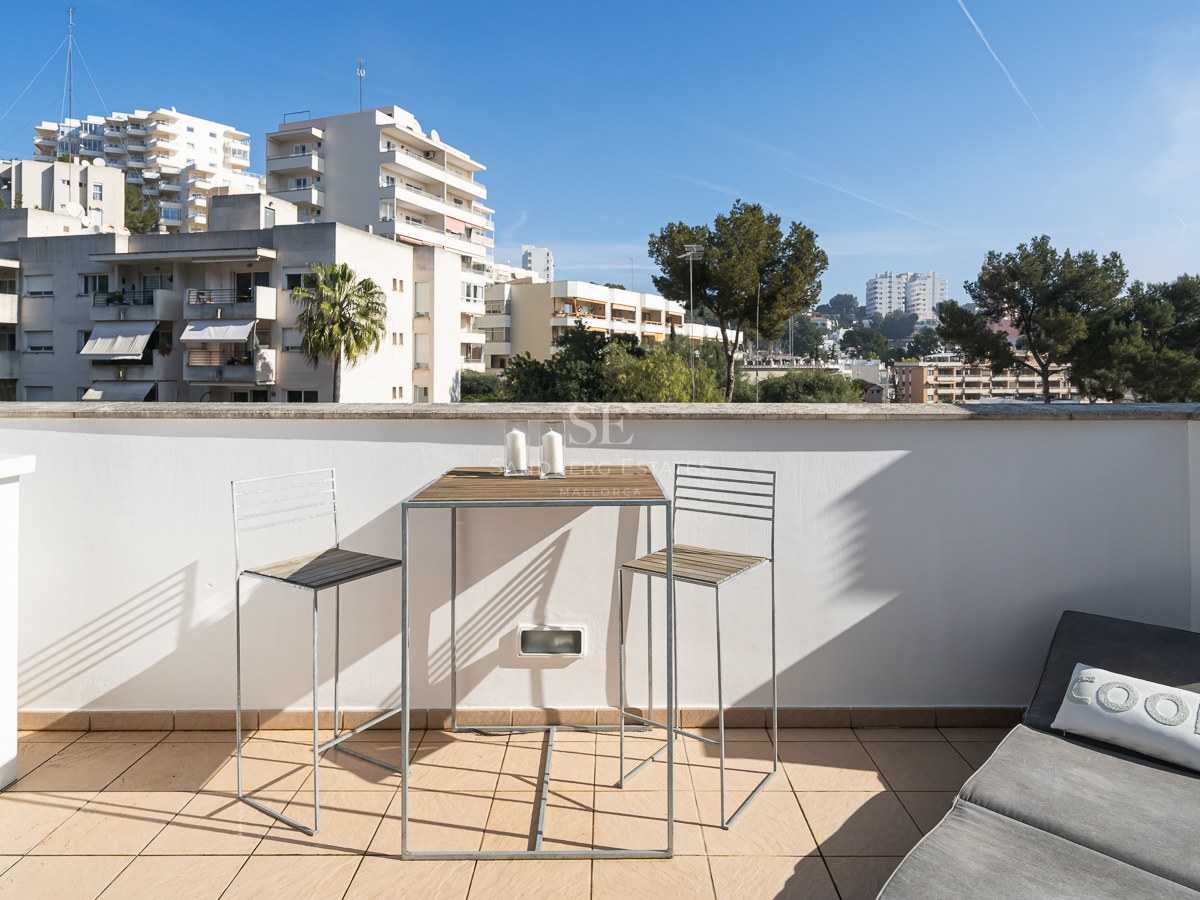 A sunlit terrace with a high-top wooden table, two bar chairs, and a grey sun lounger overlooking city buildings.