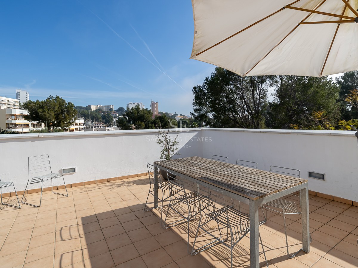 Spacious rooftop terrace with a large wooden table, wire chairs, and a white sun umbrella overlooking a city.