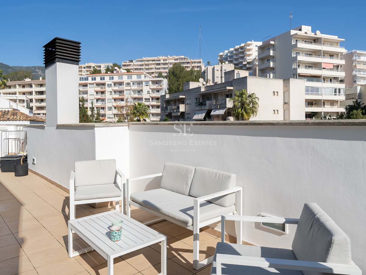 Sunlit rooftop terrace with white modern furniture, tiled floor, and views of city buildings and mountains.