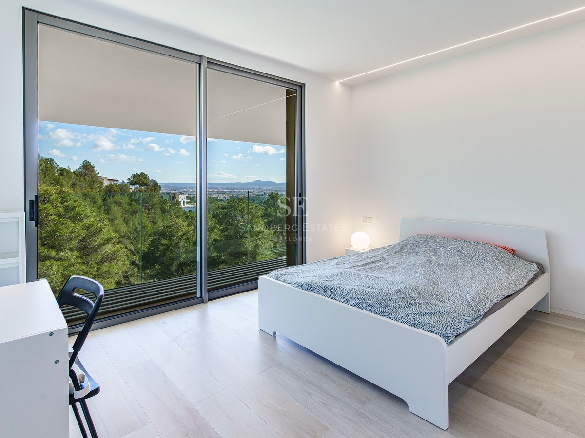 Dormitorio luminoso con cama blanca, suelo de madera clara y grandes ventanales con vistas a un bosque y montañas.