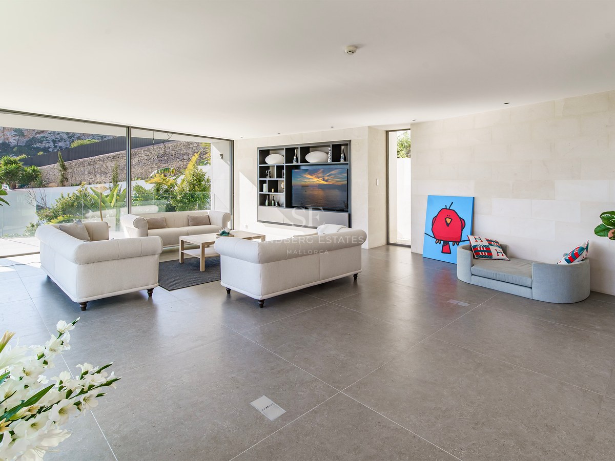 Salón moderno y luminoso con sofás beige, suelo de piedra gris y grandes ventanales hacia el jardín.