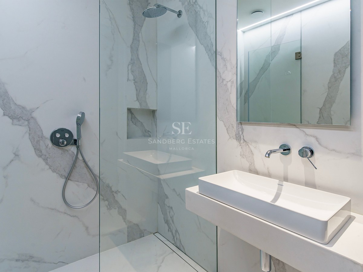 Baño moderno con azulejos efecto mármol blanco, ducha de cristal y lavabo minimalista con grifería cromada.