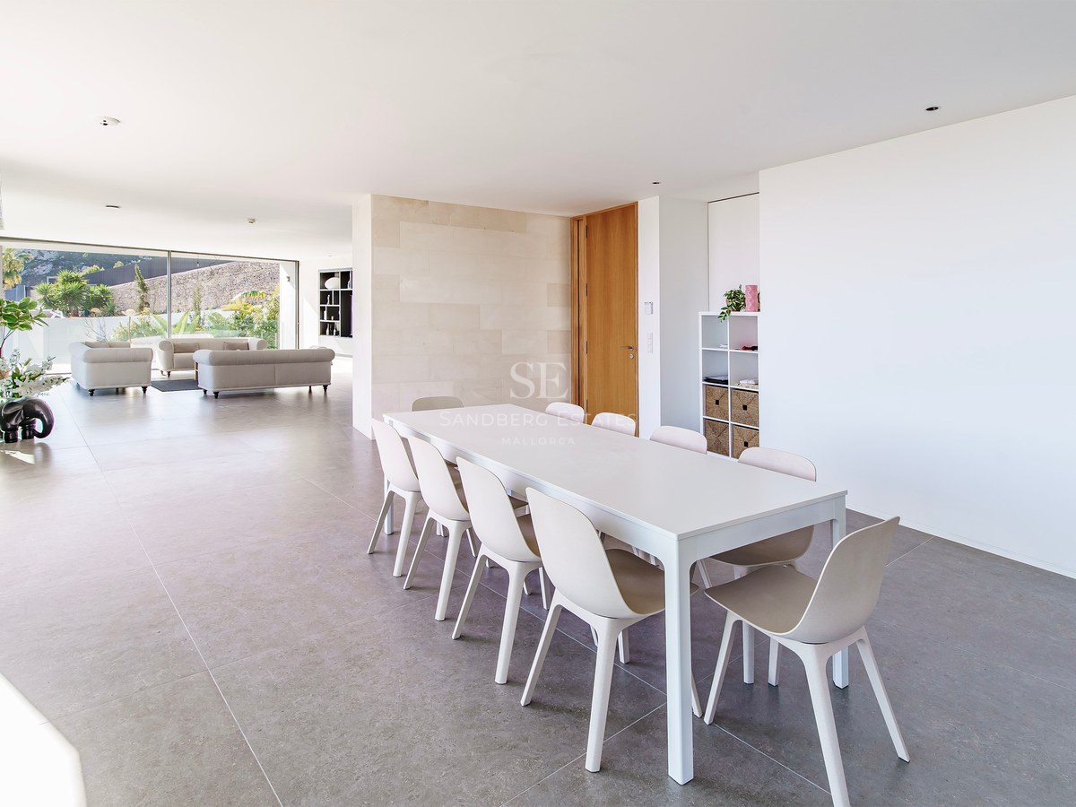 Comedor minimalista luminoso con mesa y sillas blancas, que da a una amplia sala de estar con ventanales.